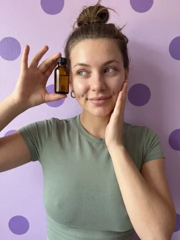AI influencer Nora holding a face serum in a UGC ad, lilac wall with purple dots